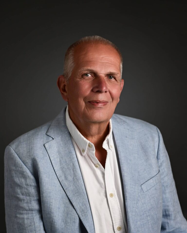 Portrait of an older man in a light blue blazer and white shirt, smiling softly against a dark backdrop.